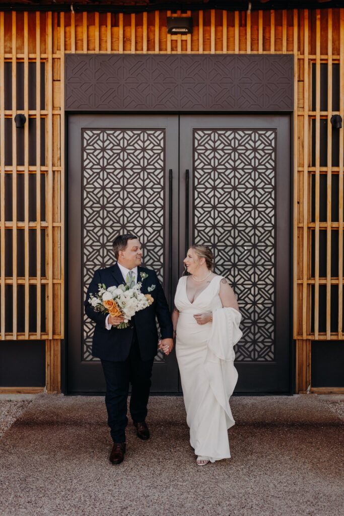 plus size bride wearing vneck crepe sheath wedding dress holding large bouquet in front of the paseo wedding venue in apache junction arizona with hsuband in dark wedding suit