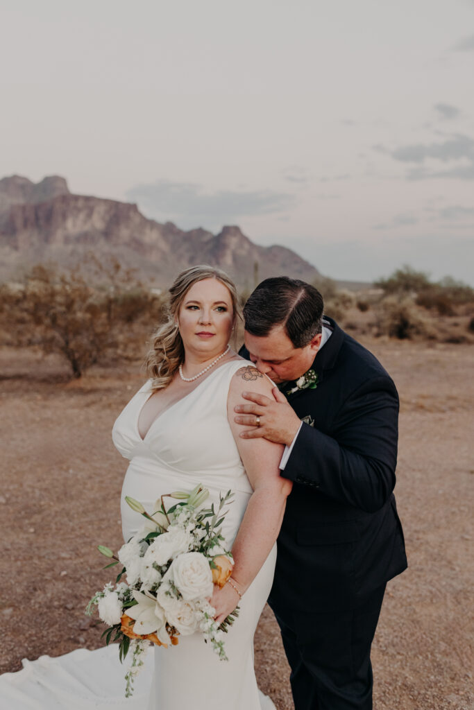 plus size bride wearing vneck crepe sheath wedding dress with husband in dark suit kissing her shoulder with superstition mountains behind them