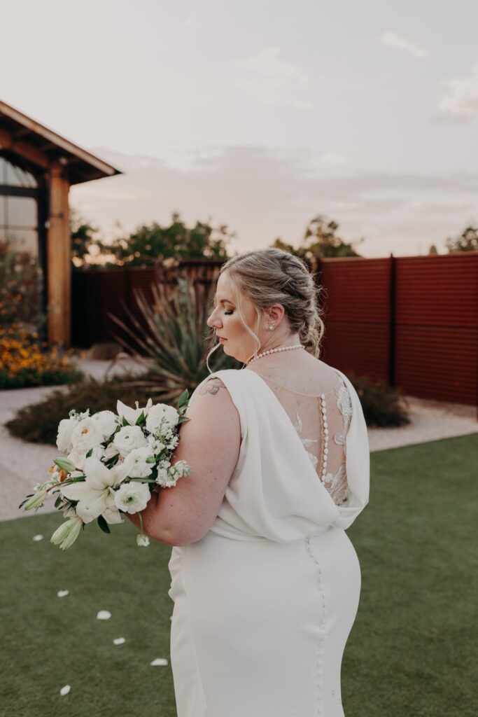 plus size bride wearing vneck crepe sheath wedding dress holding large bouquet in front of the paseo wedding venue in apache junction arizona