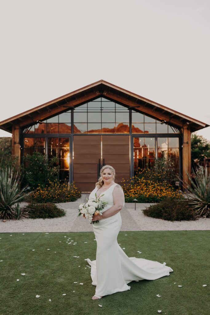 plus size bride wearing vneck crepe sheath wedding dress holding large bouquet in front of the paseo wedding venue in apache junction arizona