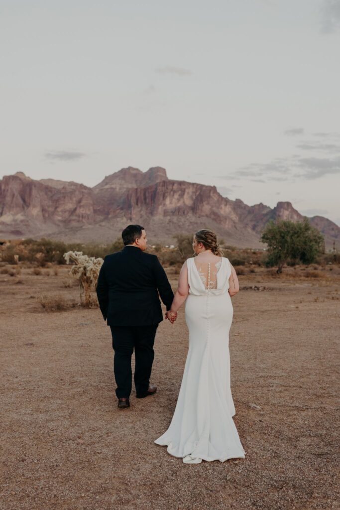 plus size bride wearing vneck crepe sheath wedding dress with husband in dark suit walking in front of superstition mountains arizona