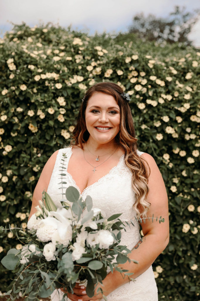 plus size bride wearing vneck lace wedding dress with greenery behind her and carrying a large wedding bouquet