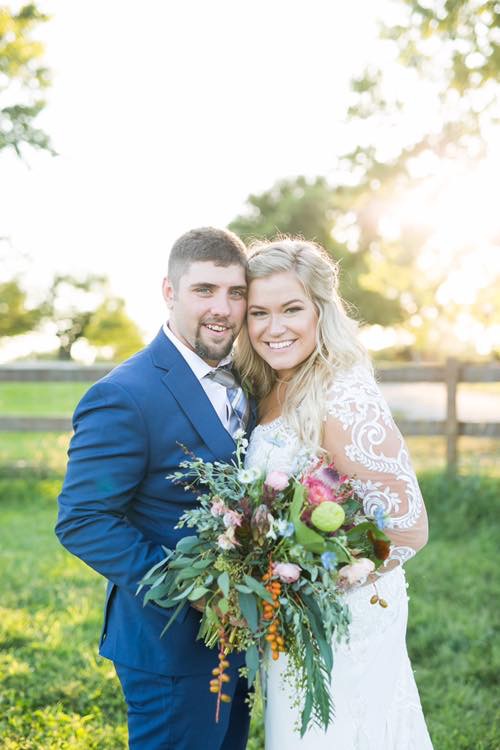 Smiling couple on wedding day with rustic bouquet with embroidered long sleeve gown and navy tuxedo
