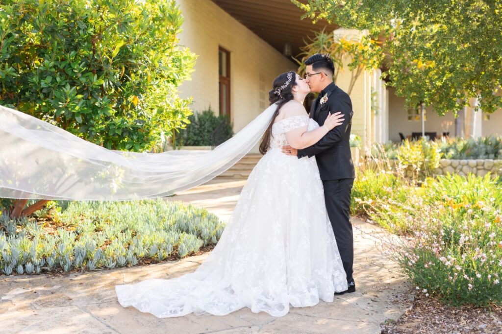 bride wearing lace ballgown with long veil kissing groom in black tux