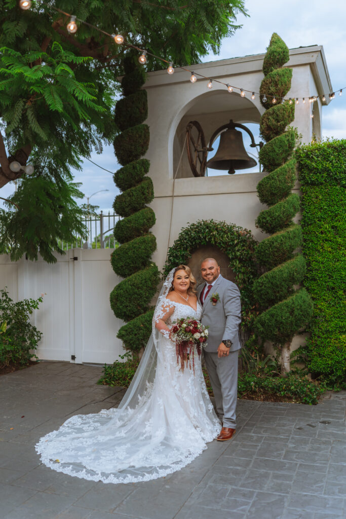 plus size bride wearing long sleeve lace fitted wedding dress with long dramatic lace train with groom in gray suit outdoor wedding