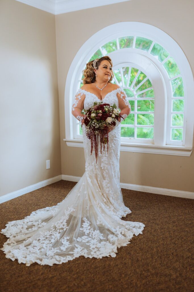 plus size bride wearing long sleeve lace fitted wedding dress with long dramatic lace train holding red bouquet