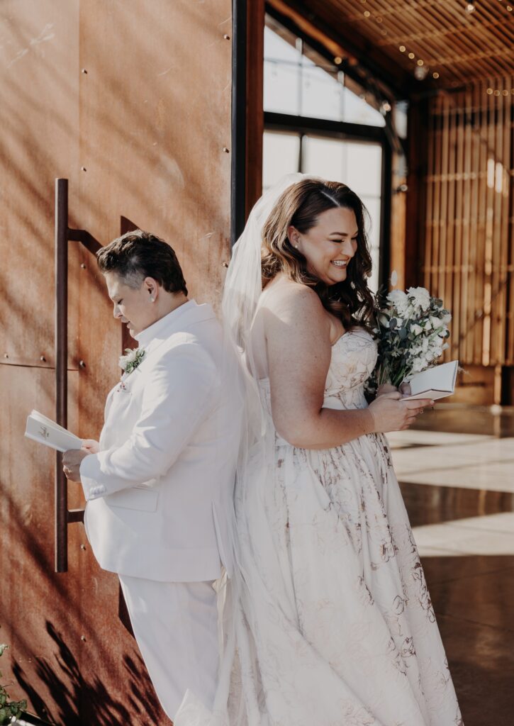 bride in white suit and bride in unique wedding dress reading wedding vows inside barn chandler arizona